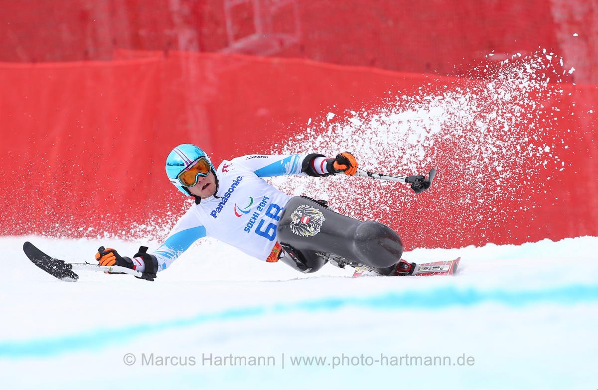 Sit skier Roman Rabl rounds a turn very low, at speed with snow spraying behind him and outriggers outstretched.