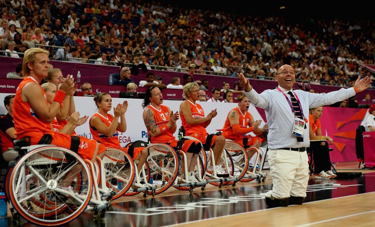 Female wheelchair basketball players lined up, cheering during a game with an amputee man in a suit standing in front of them