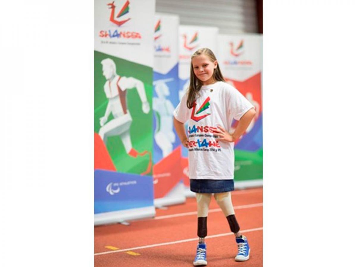 A young amputee girl smiles to the camera, wearing a white t-shirt with a Swansea 2014 imprint