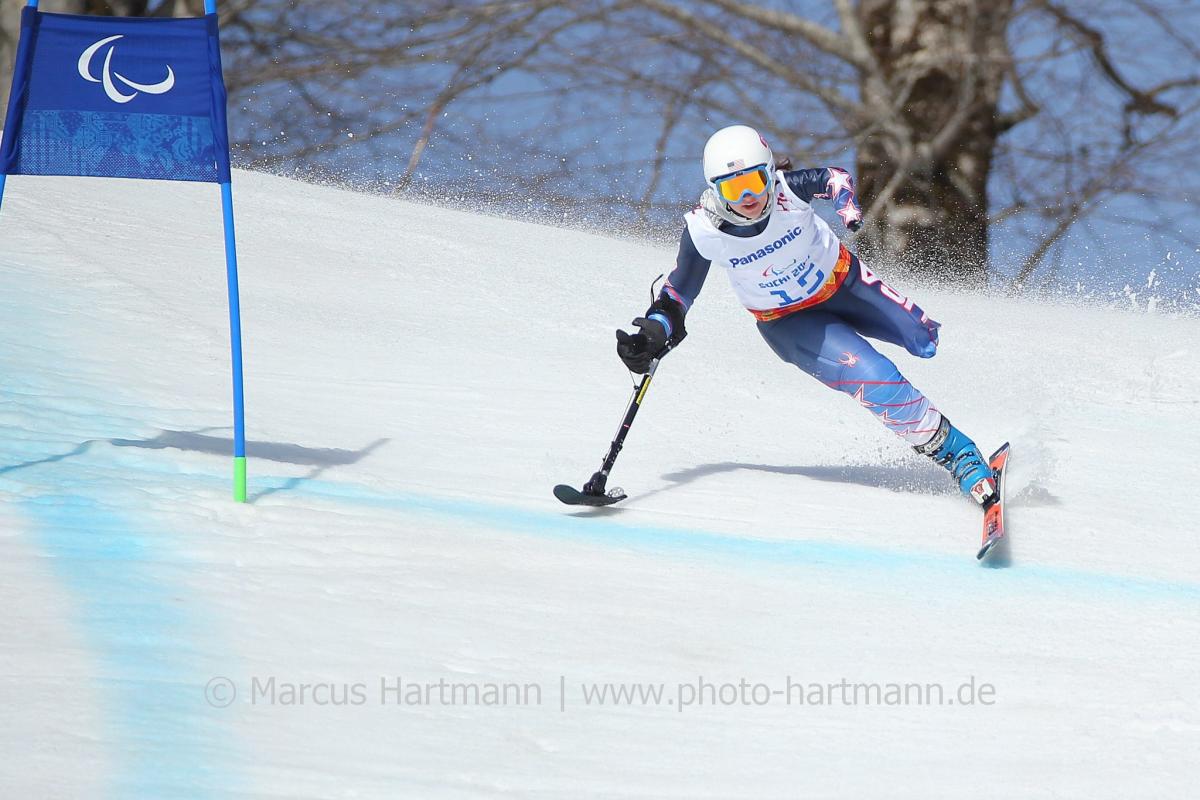 Skier with one leg skis at speed past a gate with her outrigger outstretched