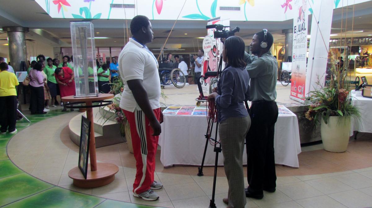 Team of a camera man and a women holding a microphone stand opposite of a man in sport dress. They seem to be preparing an interview. Background: A stage with a wheelchair on it