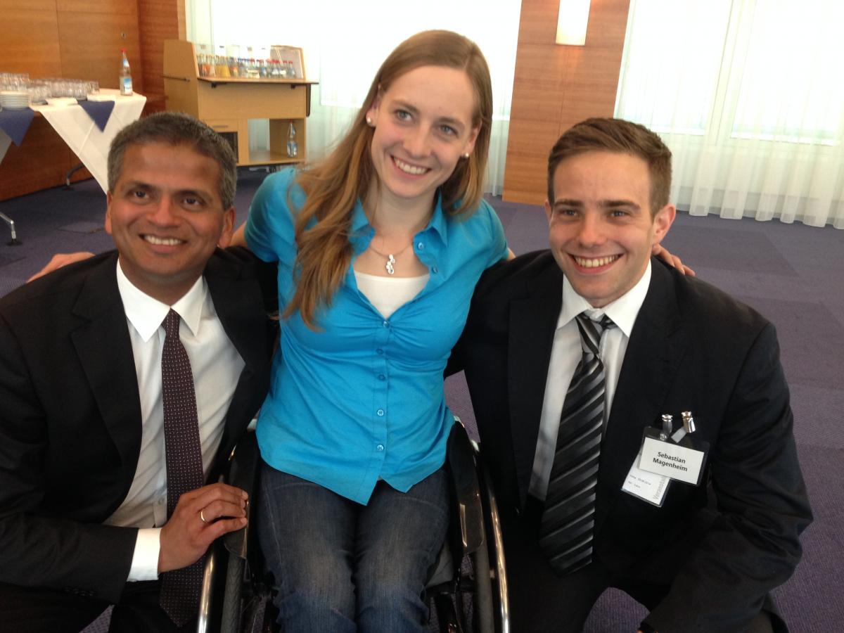 Women in wheelchair smiling, poses surrounded by two men in suits who perch next to he