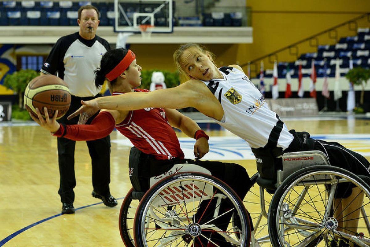 Two female basketball players in wheelchairs fight for the ball. A referee watches them from behind