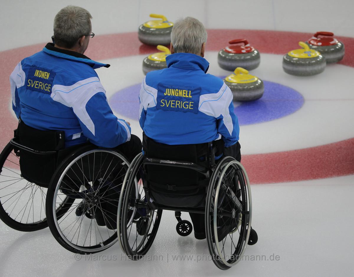 Two men in wheelchair on ice, looking at some curling stones.