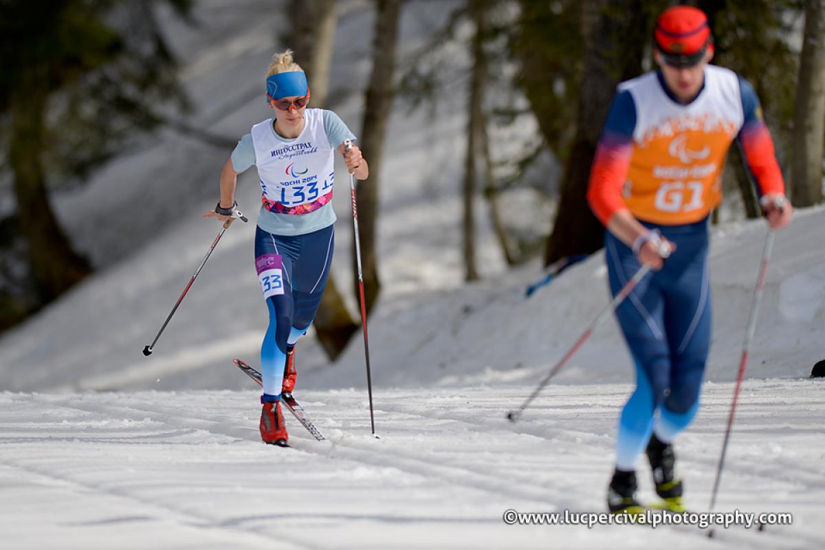 Female cross country skier behind a male cross country skier with orange bib on a track