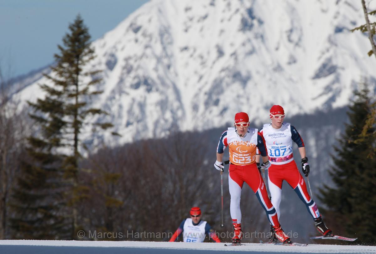 Wide shot of two Nordic skiers skating close together on the crest of the hill, with one skier a short distance behind. Big mountain in background