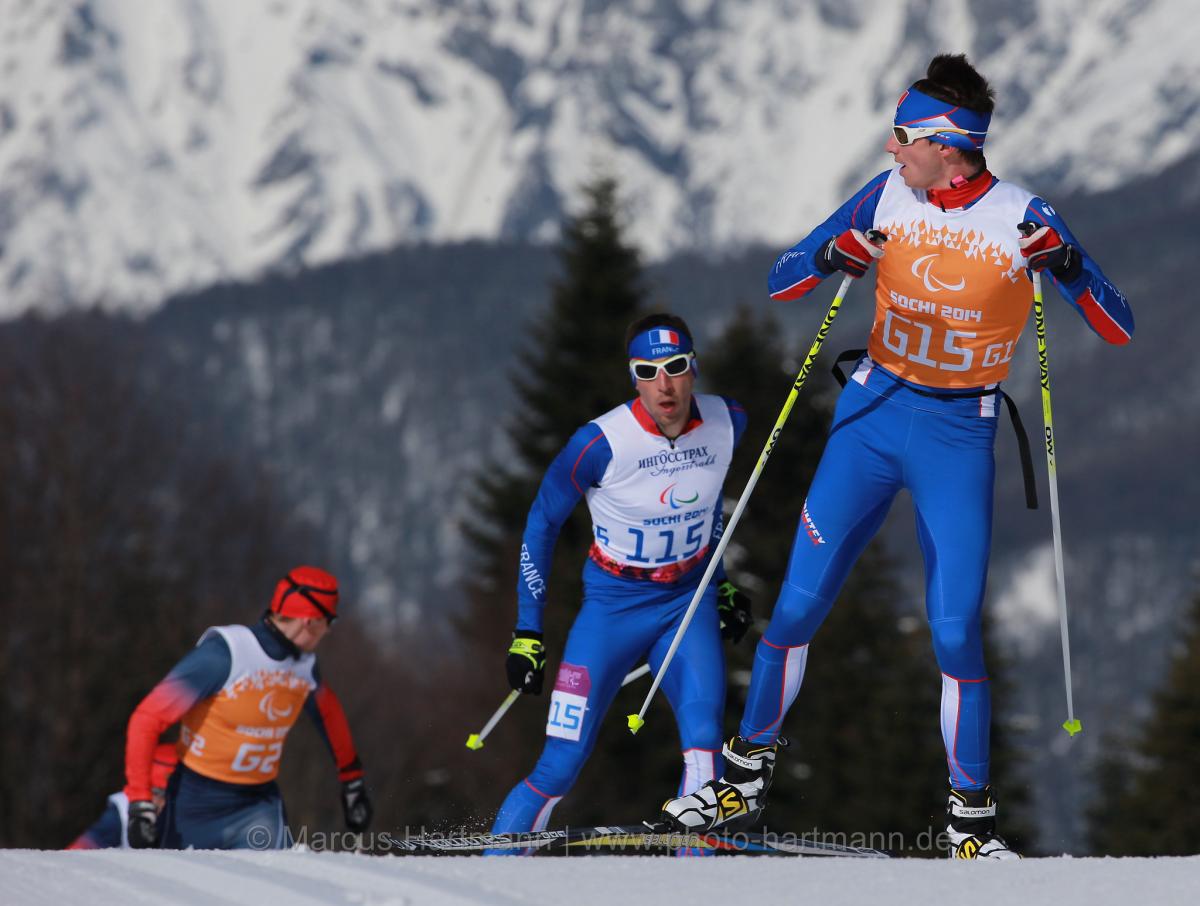 A Nordic skier looks behind him at two other skiers approaching the crest of the hill, close behind him. Mountains are in the background.