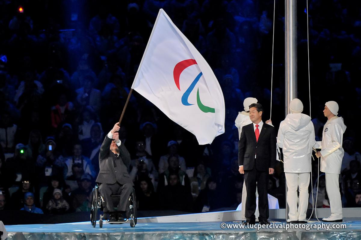 Man in wheelchair on a stage waving a huge flag with the Paralympic symbol on it, handing it over to another person.