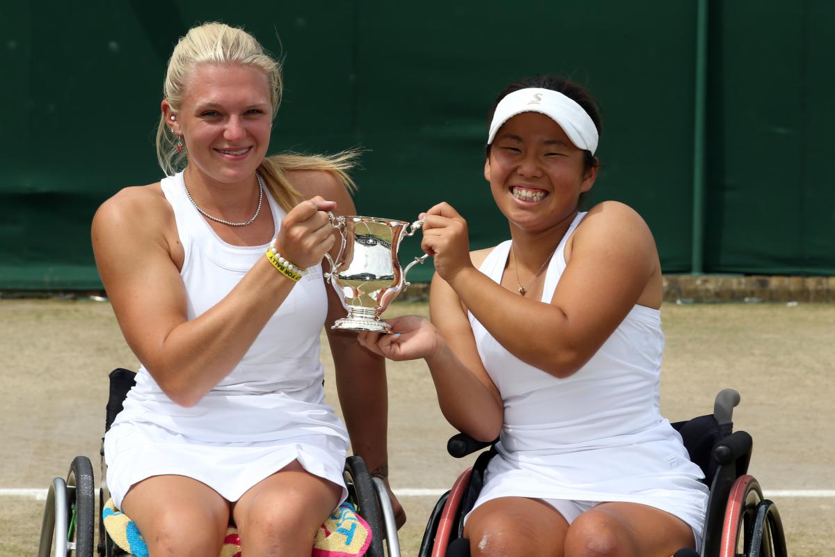 Two women in wheelchairs on a tennis court hold a trophy and smile to the camera.