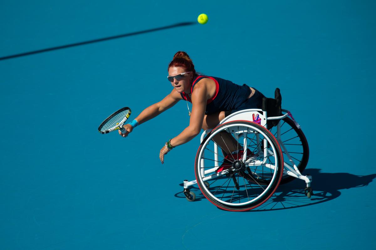 Woman in a wheelchair on a blue tennis court trying to reach a ball