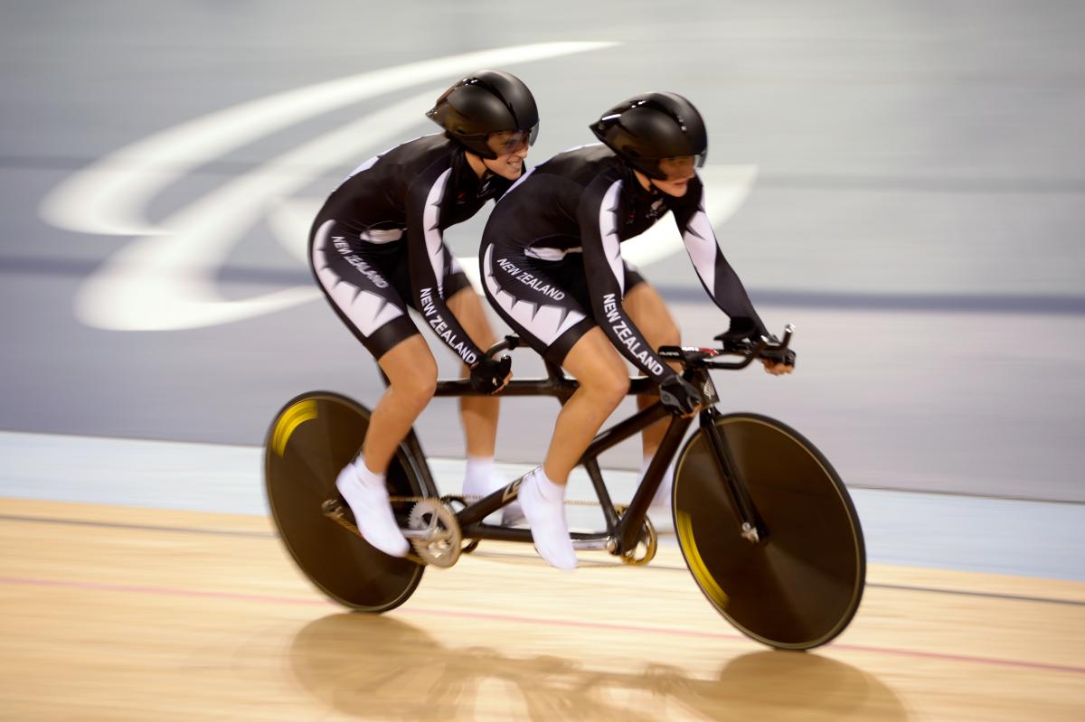 Two female cyclists in black racing suits on a tandem, competing at the London 2012 Paralympic Games.