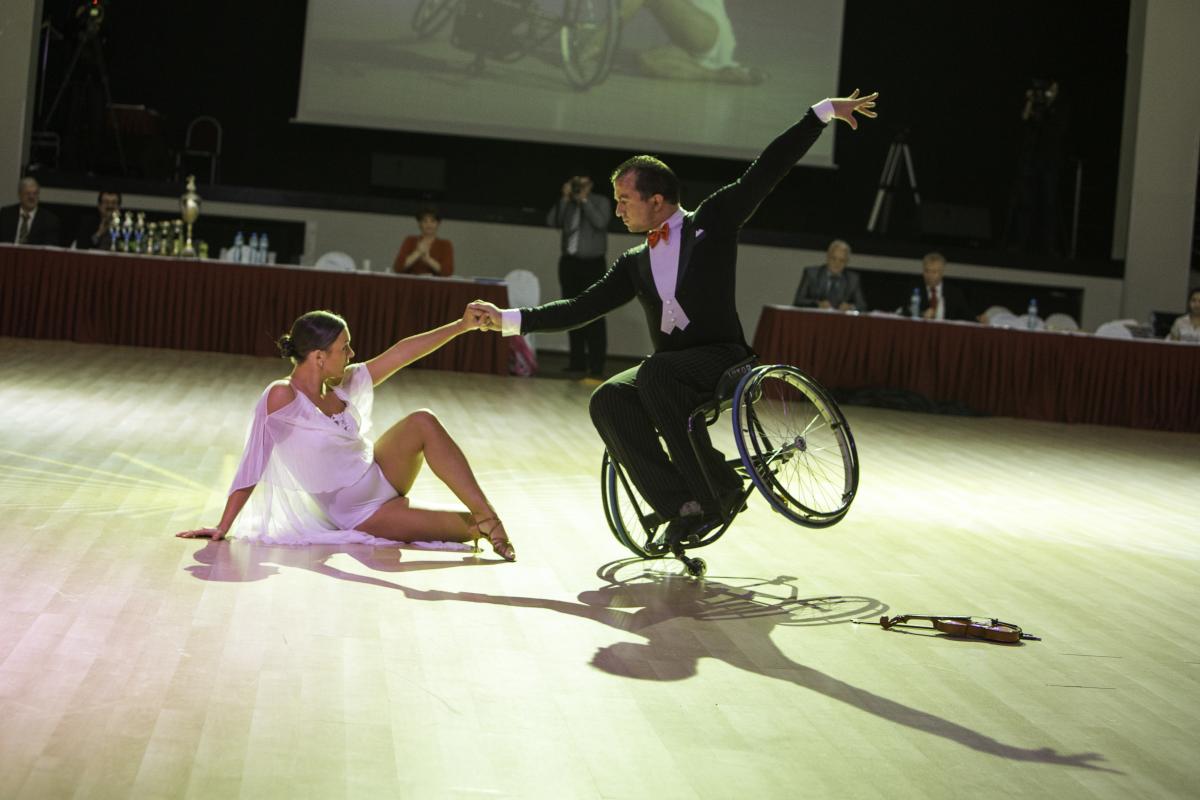 Couple on the dance floow. He is in a wheelchar, she is sitting on the floor, both pose