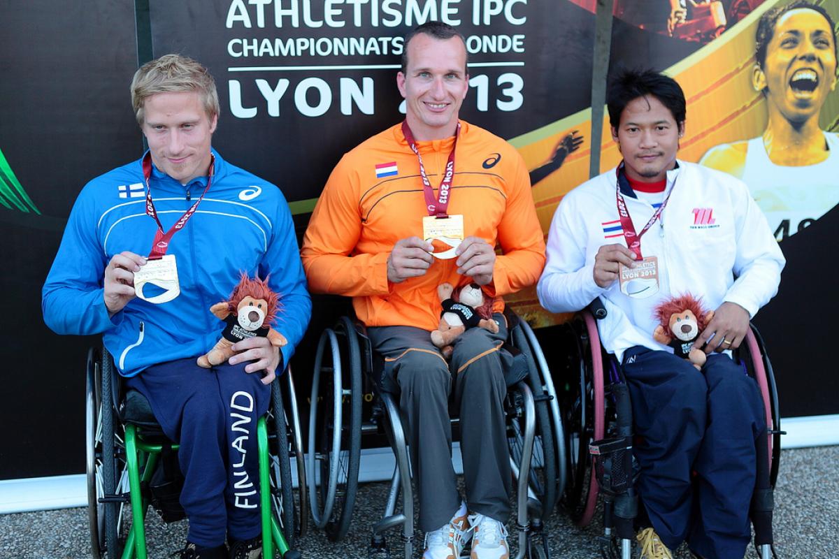Three wheelchair athletes pose with their medals in front of a Lyon branded background