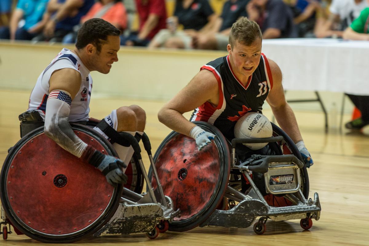 A player wearing blue seated in a wheelchair with a white ball on his lap about to collide with an opponent wearing white.