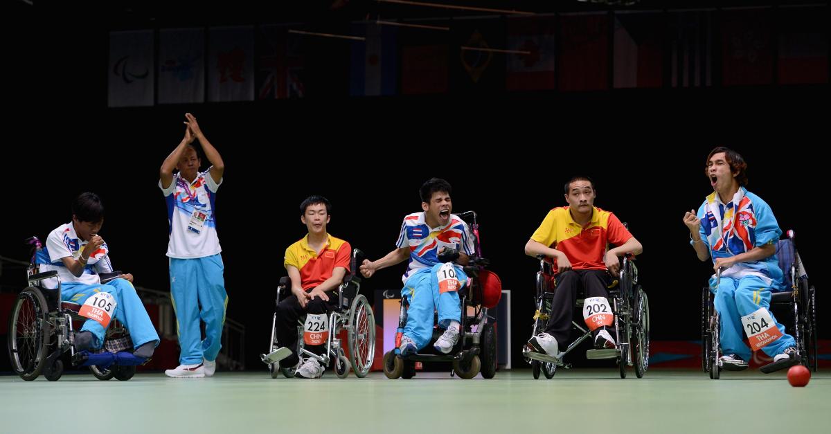 Four men in wheelchairs and one man standing on a field of play with dark background. Three men celebrating