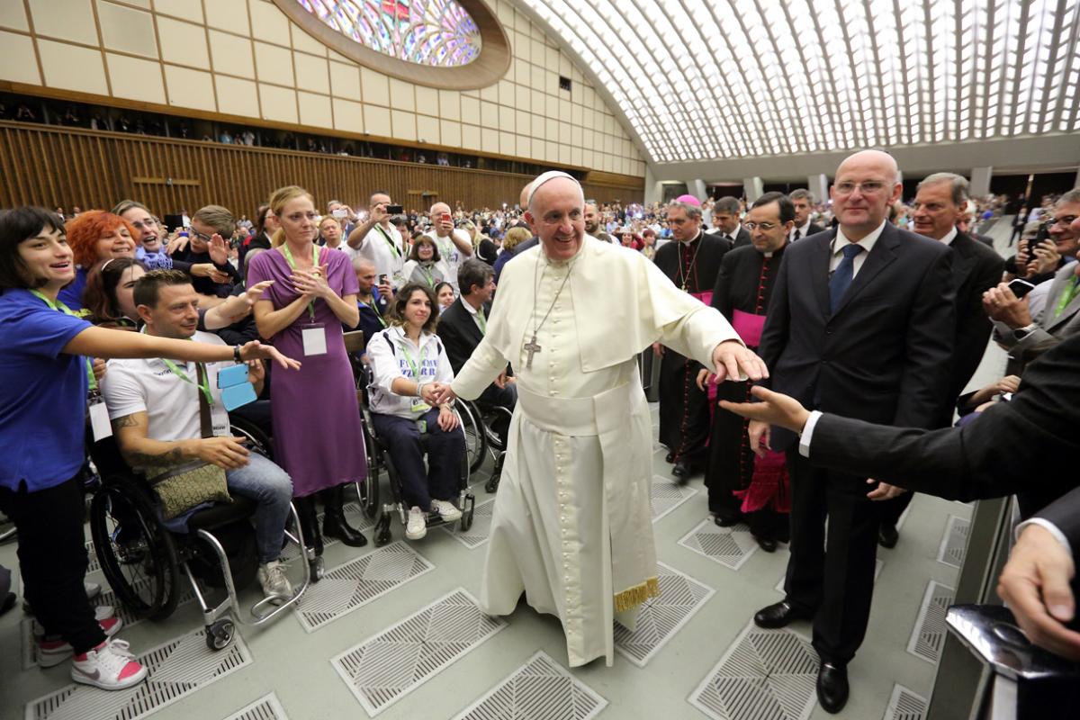 Man in white robe walks through a crowd of people, some of them in wheelchairs