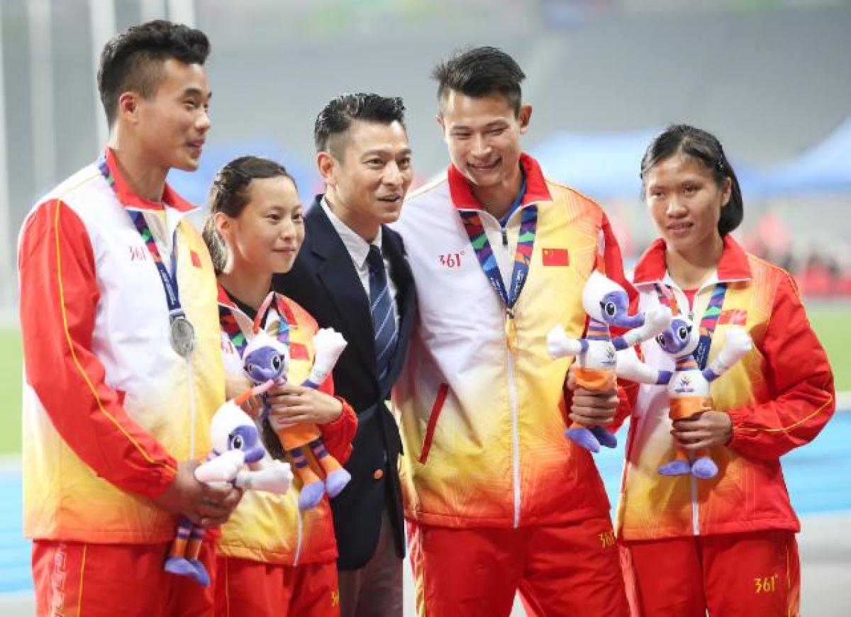 Man in dark suit stands between some athletes on the podium 