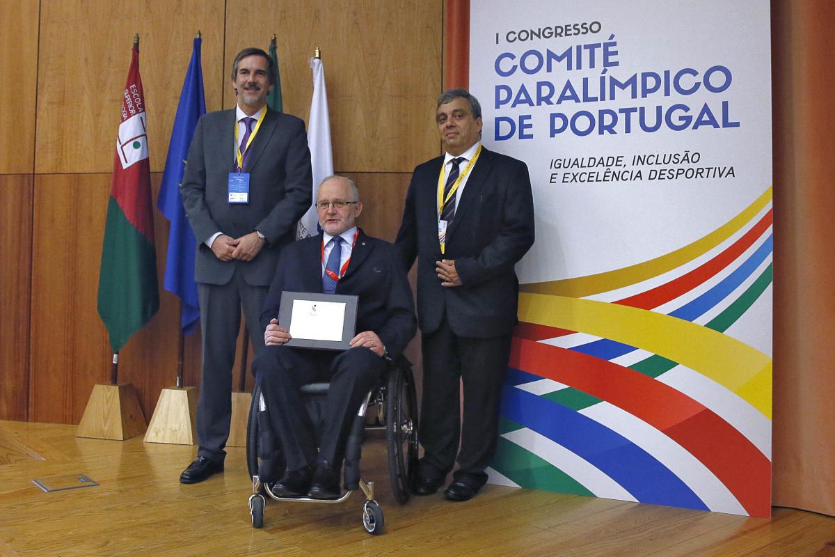 Three men (one in a wheelchair) pose in front of a wooden wall and some flags