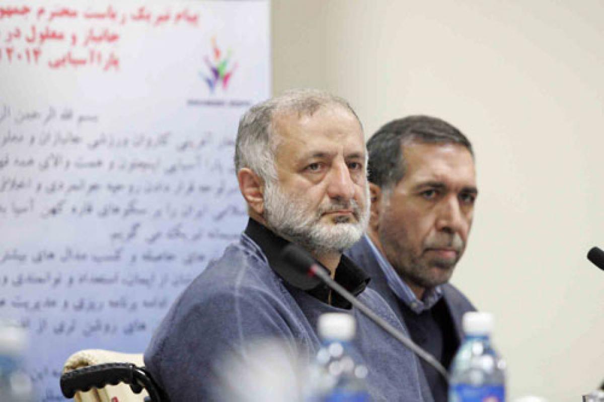 Iran NOC President Upper bodies of two men sitting behind a table, listening to something.