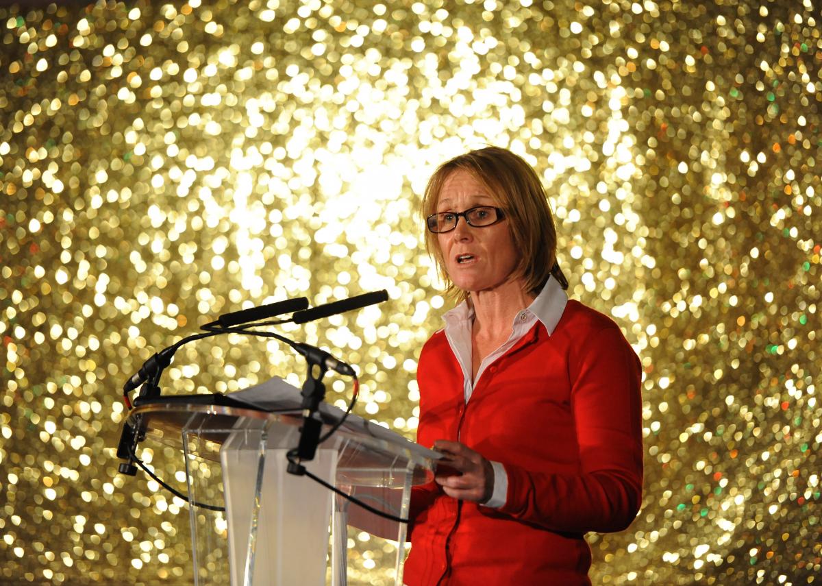 woman in red standing on stage in front of a gold background and addressing the crowd