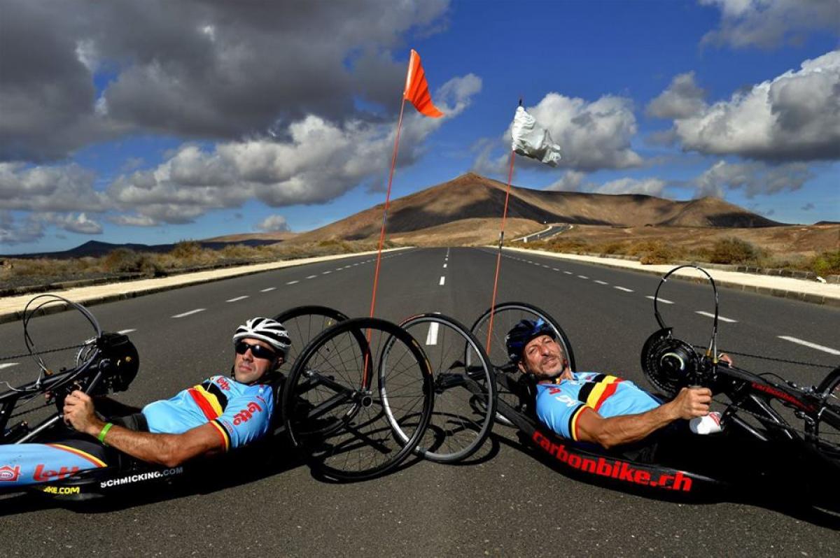 Wearing blue lycra outfits, two men lie in their handcycles on an open road with hills in the background.