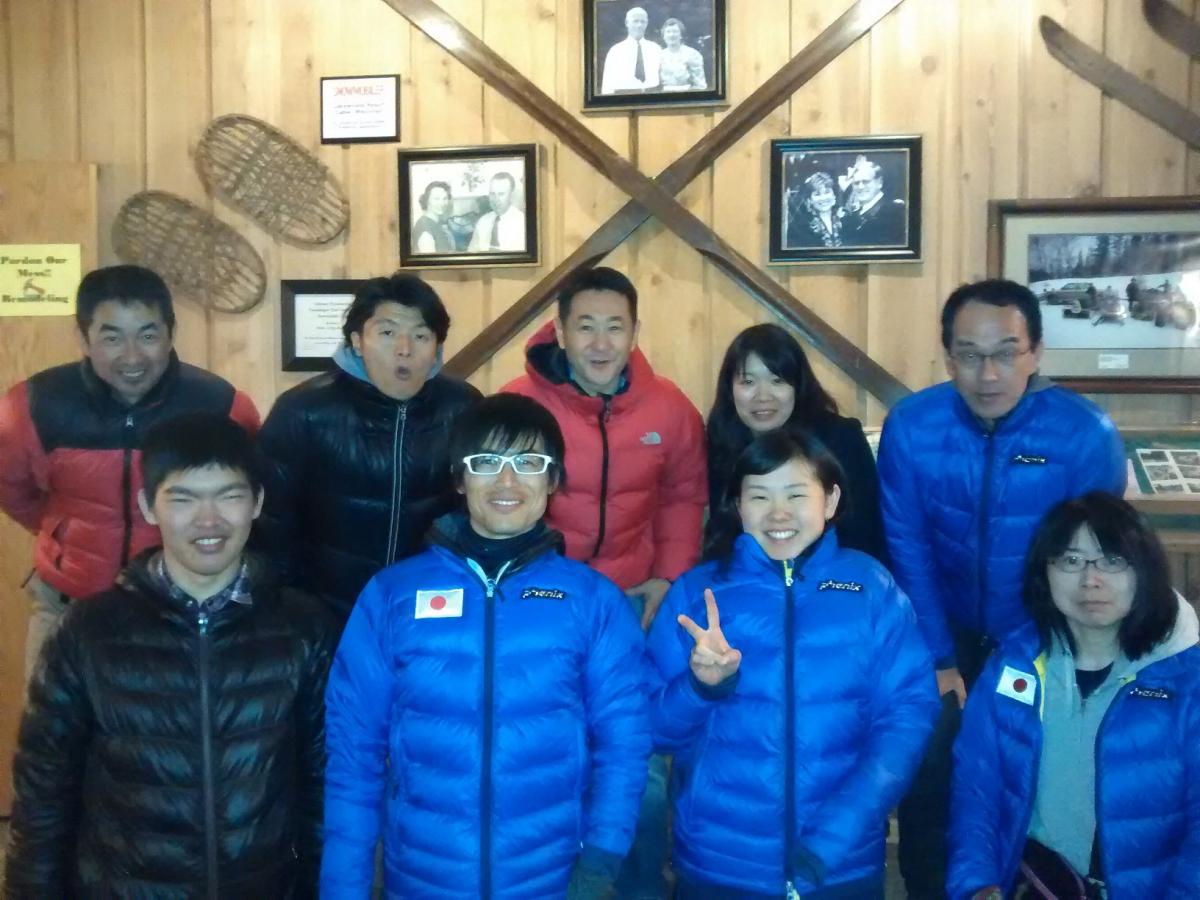 Group shot of six people in front of a wooden wall, smiling to the camera