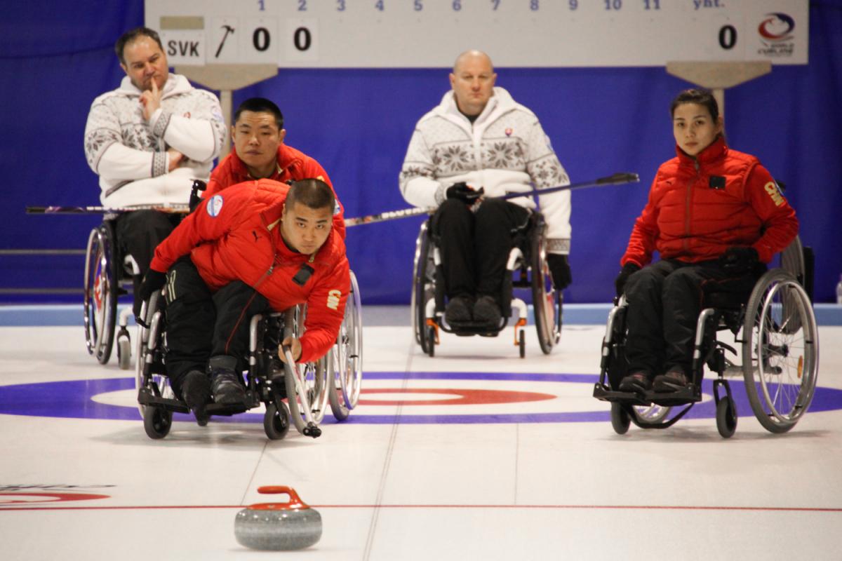 Chinese man in a wheelchair using a stick to throw a curling stone.