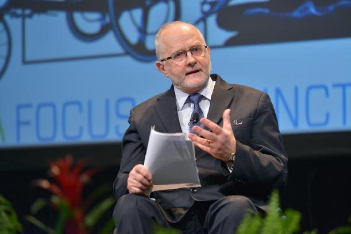 A man seated holding papers in one hand and using his other hand to express his feelings whilst giving a talk.