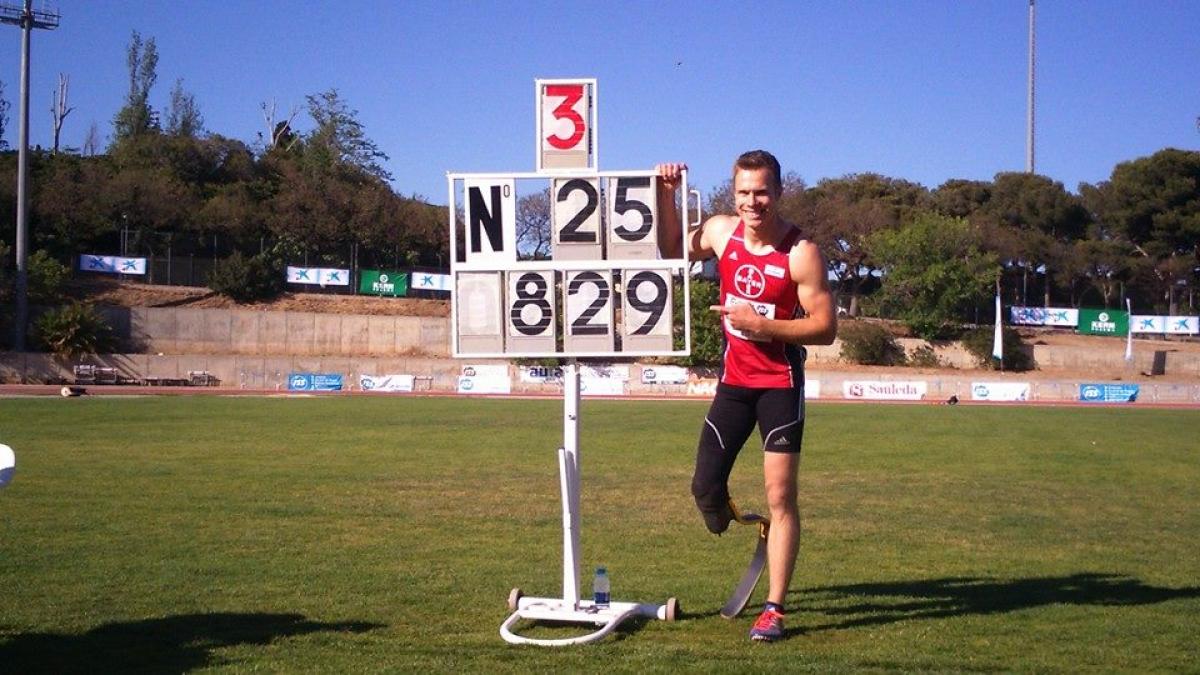 Man with prosthetic leg in a stadium, posing next to a sign that shows the number 8.29m