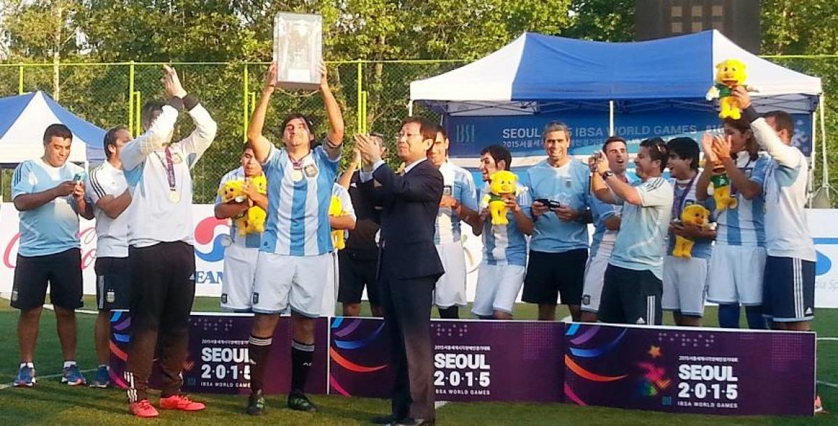 2015 IBSA World Games football-5 winner Argentina Group of men standing behind a banner holding a cup in the air.