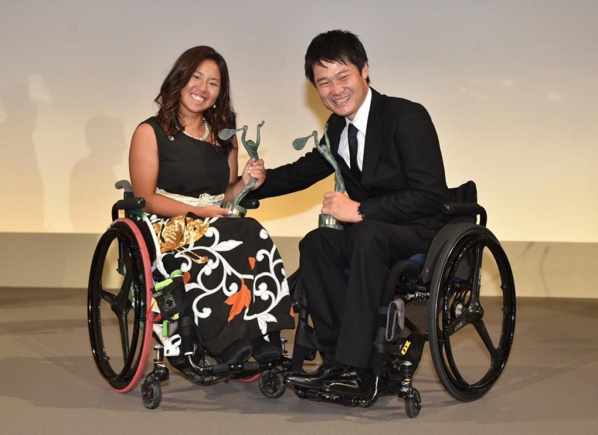Man and woman in wheechairs holding trophies and smiling to the camera