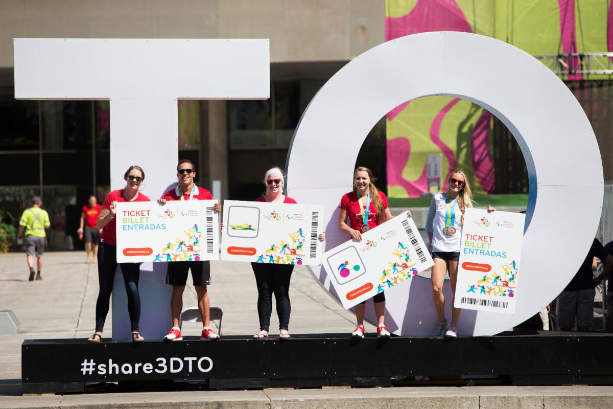 Five persons with big tickets in their hands pose in front of a big sign with the letters TO