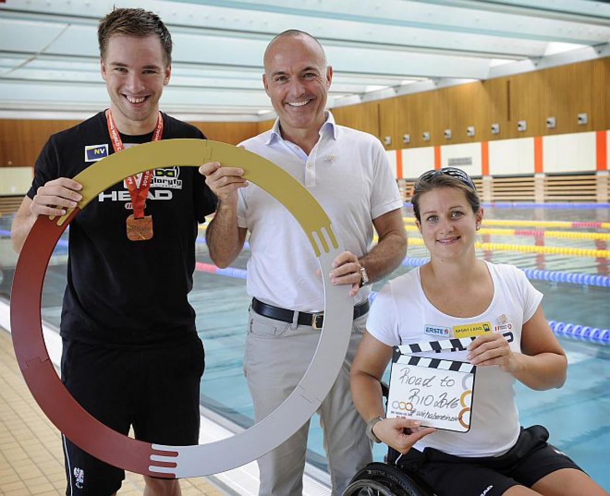 Two men and a woman in a wheelchair pose in front of a swimming pool.