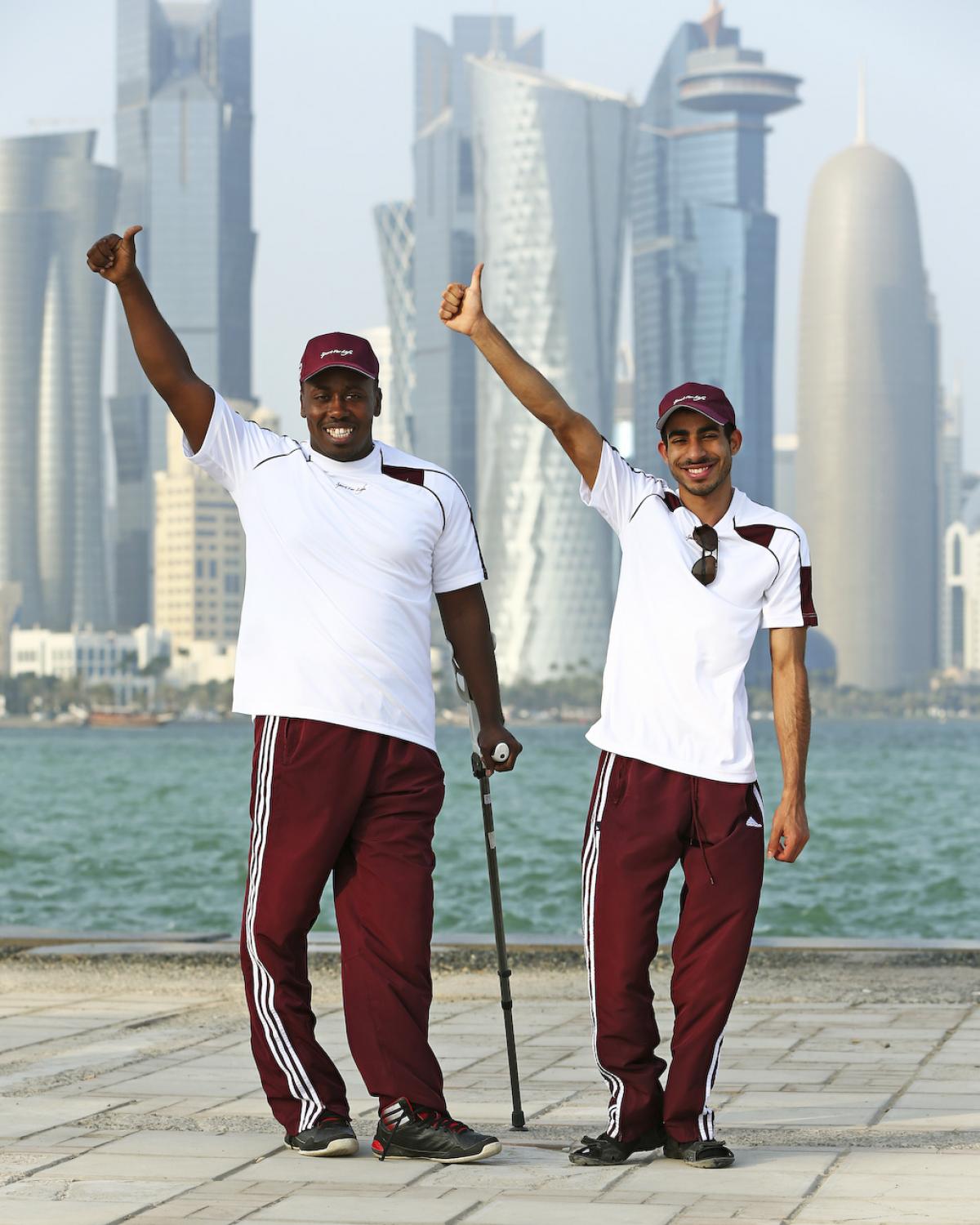 Two men in training suits in front of a skyline, waving