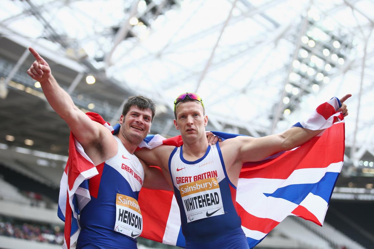 Two men in British racing vests hold a union jack behind them as they celebrate victory,