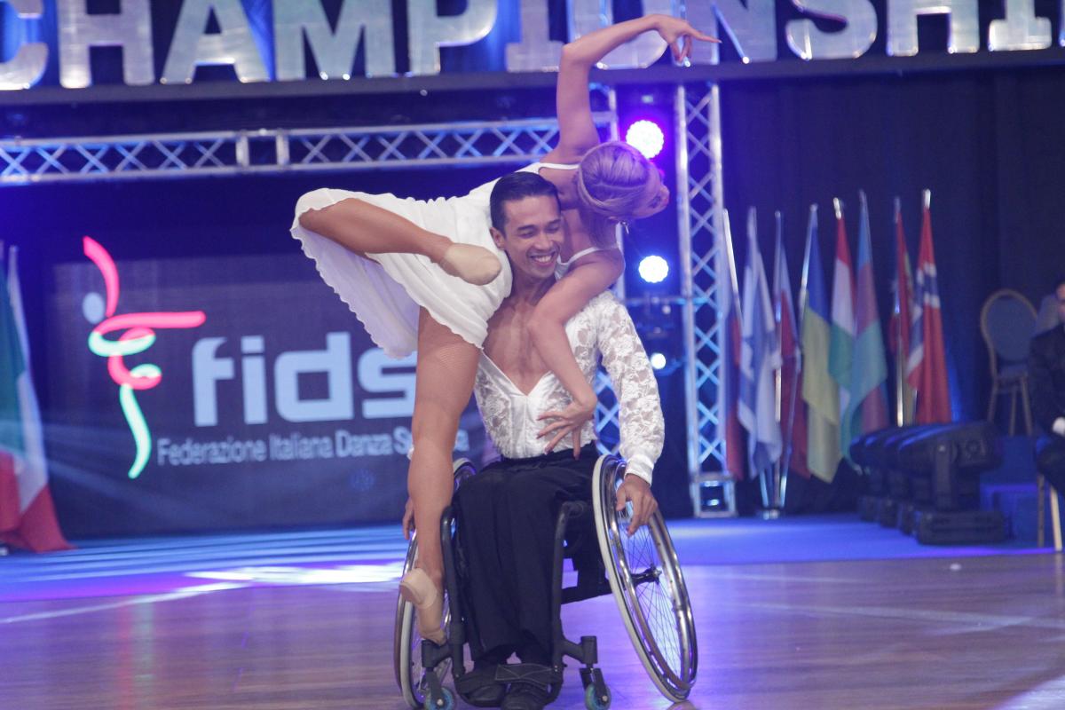 Man in wheelchair holds woman in air while spinning wheelchair.