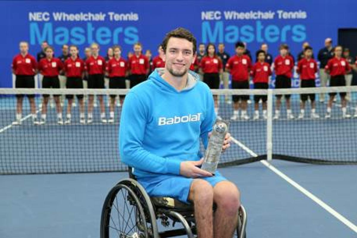 Man in wheelchair on a tennis court, showing a trophy to the camera