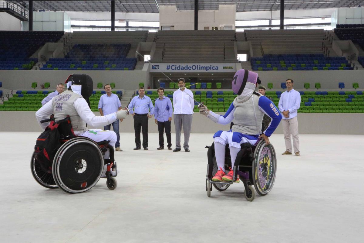 Two wheelchair fencers cross swords in empty arena, some people watching from behind