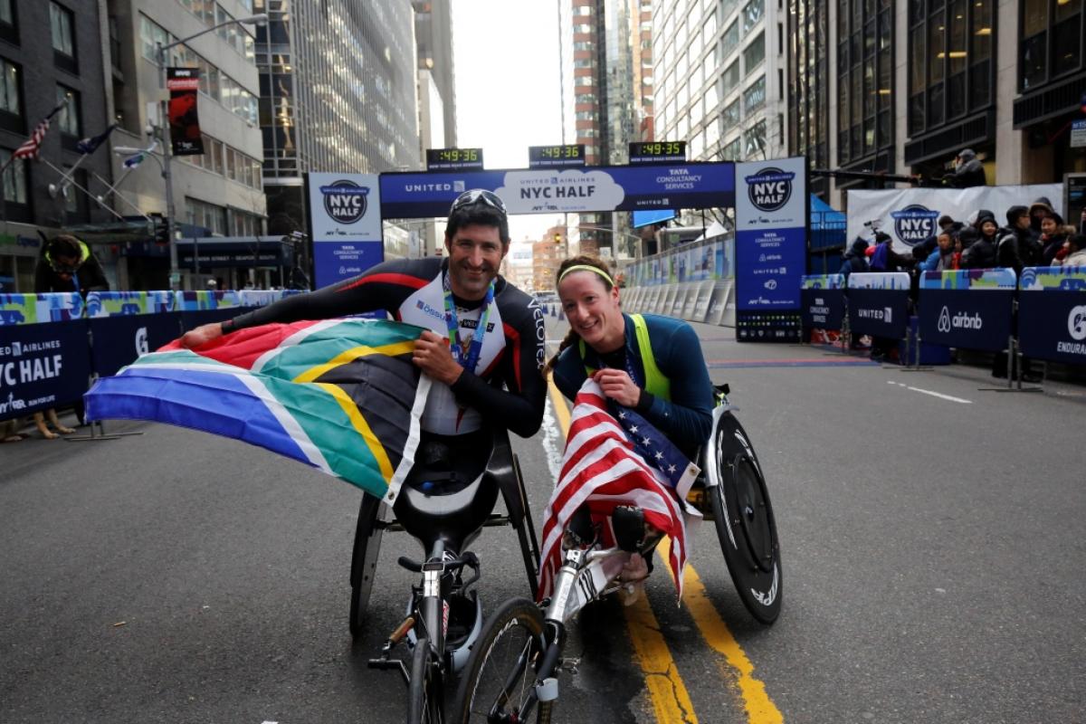 A man and a woman in racing wheelchairs, posing and showing an American and a South African flag
