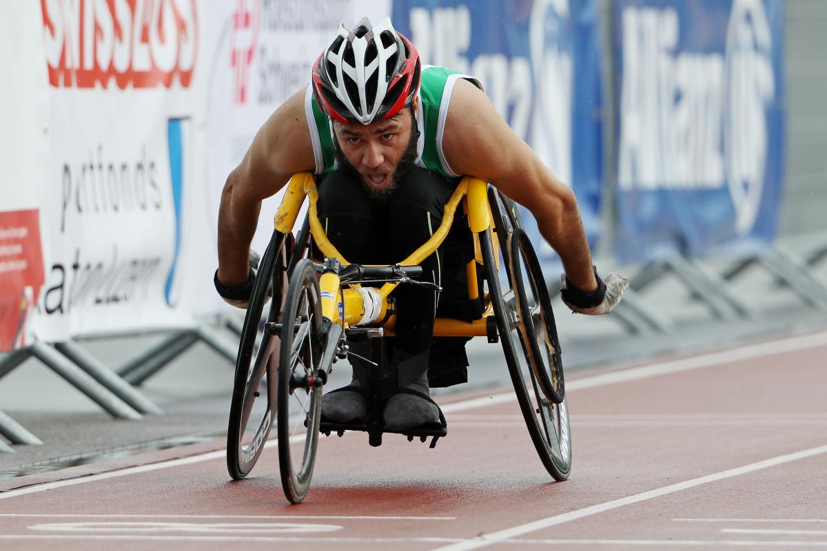 Mohamed Berrahal A man in a yellow racing chair pushes forward in the outside lane of a race.