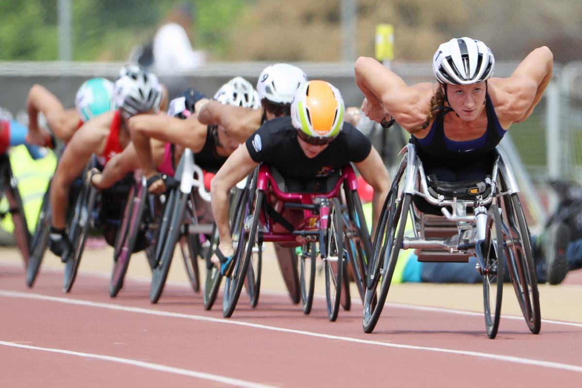 A wheelchair racer leads a line of competitors on a red track.