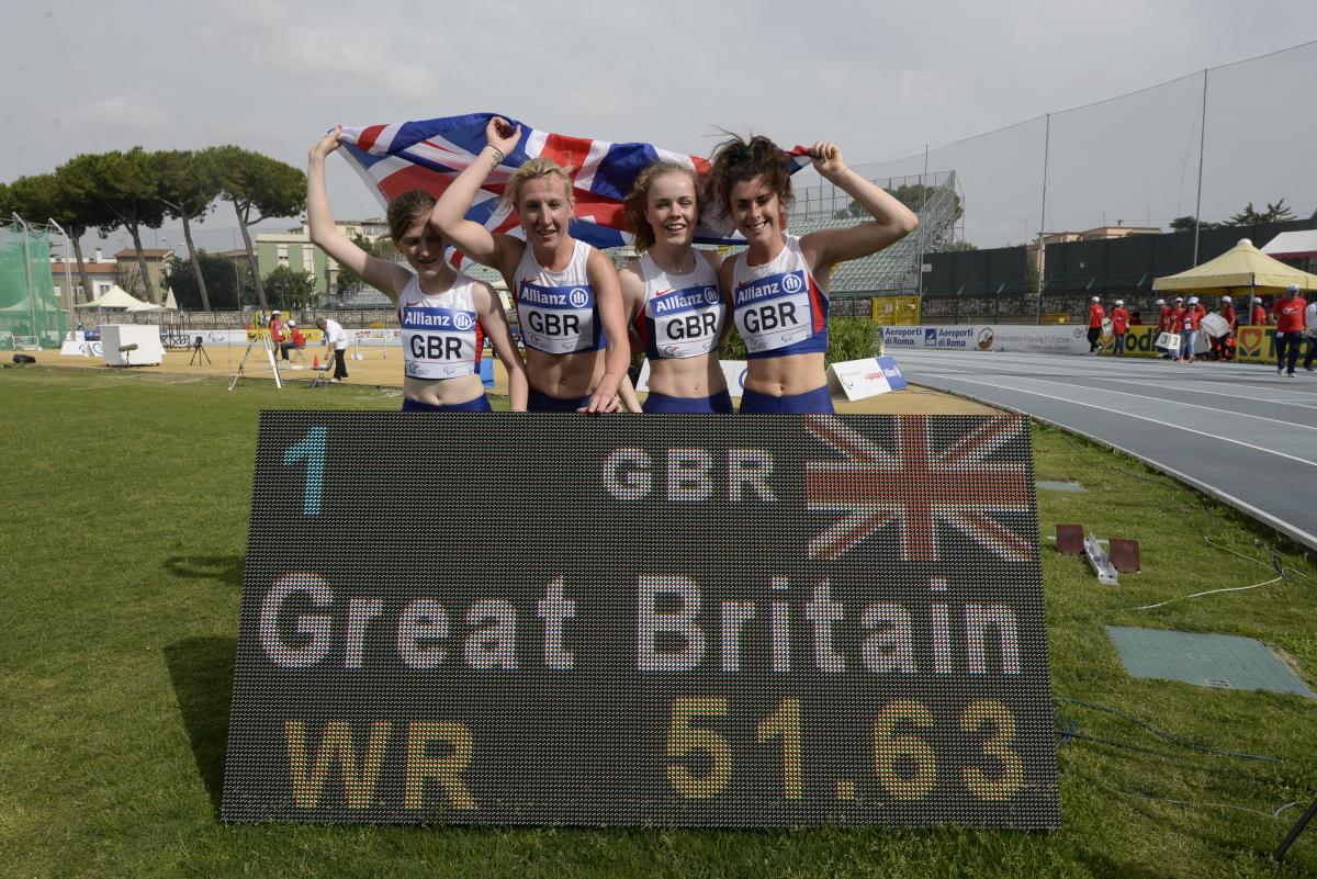 Four girls with British flag post behind a scoreboard