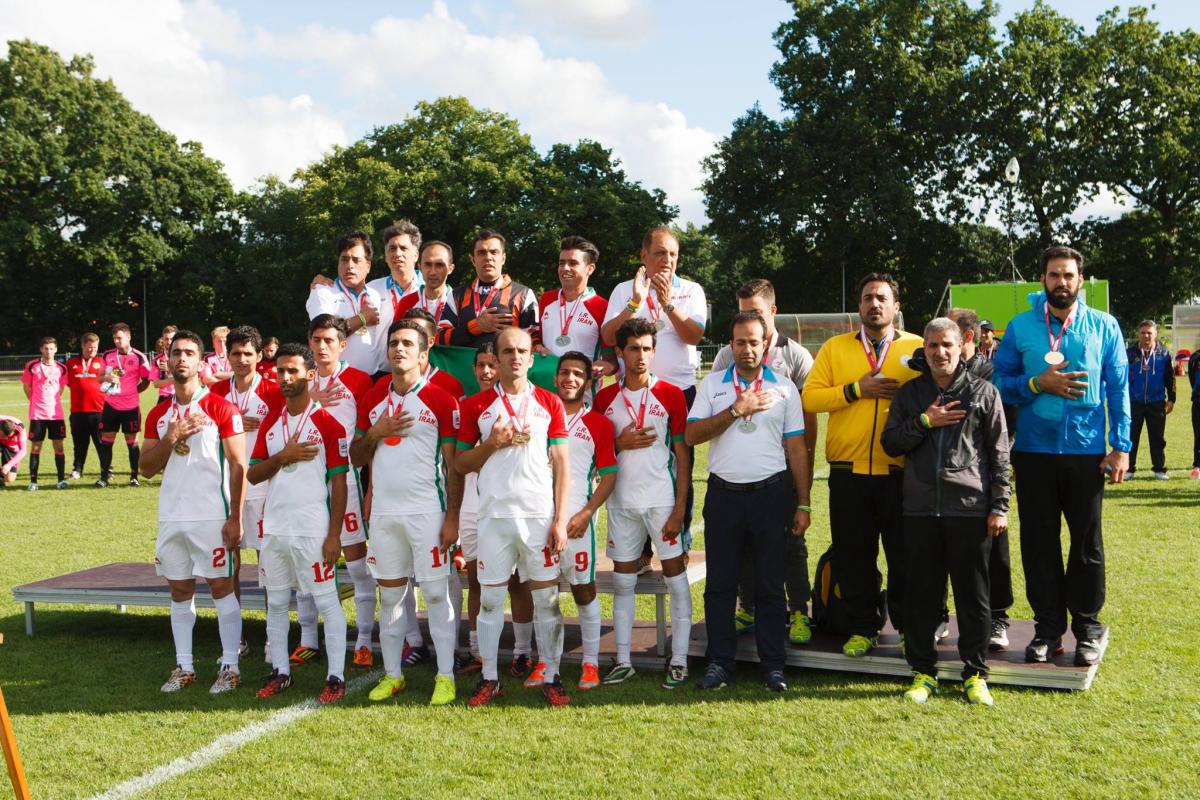 Iranian football 7-a-side team and their staff stand together, put their right hand ver their chests.  