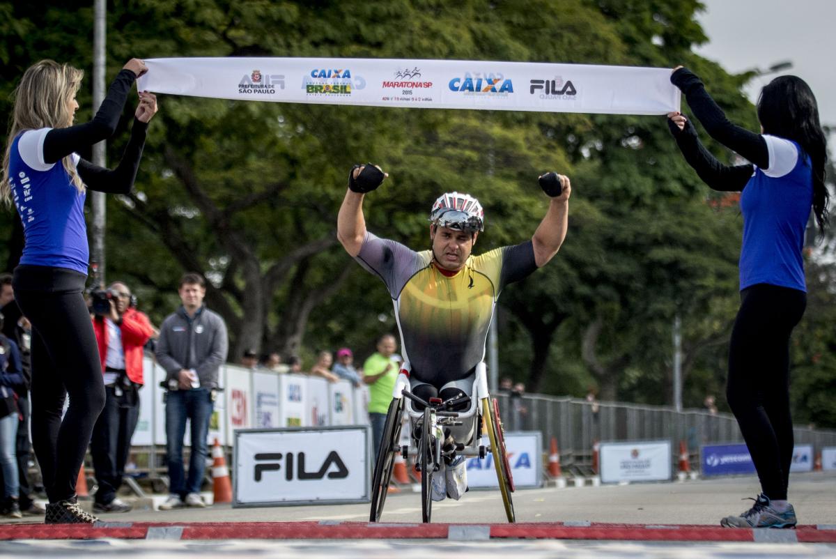 Man in wheelchair raises both arms as he crosses a finishline. 