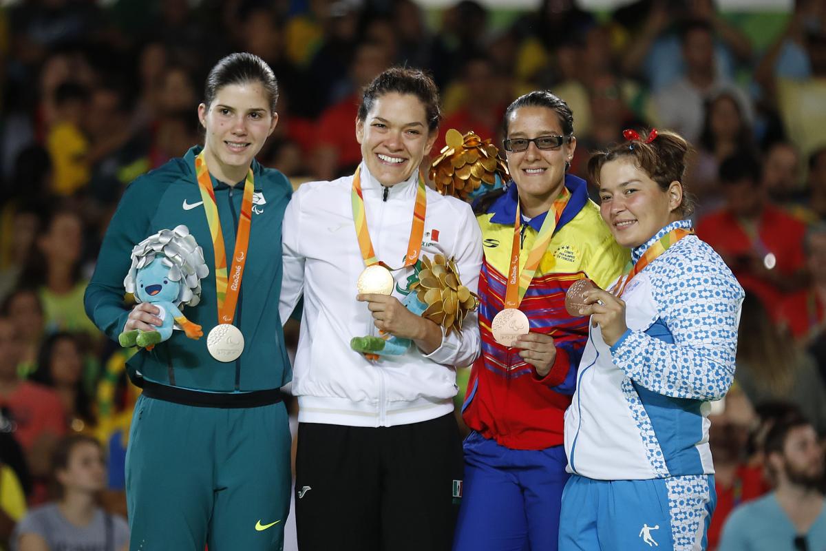 Four women in training suits on a podium, showing medals and mascots