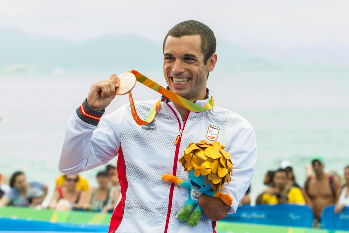 man stands on podium holding up a medal and a mascot