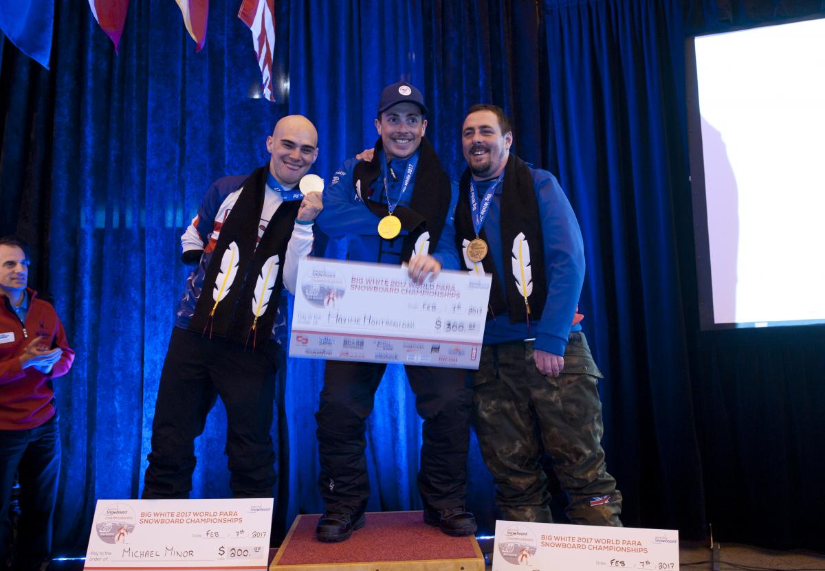 Three men on a podium holding giant checks and their medals
