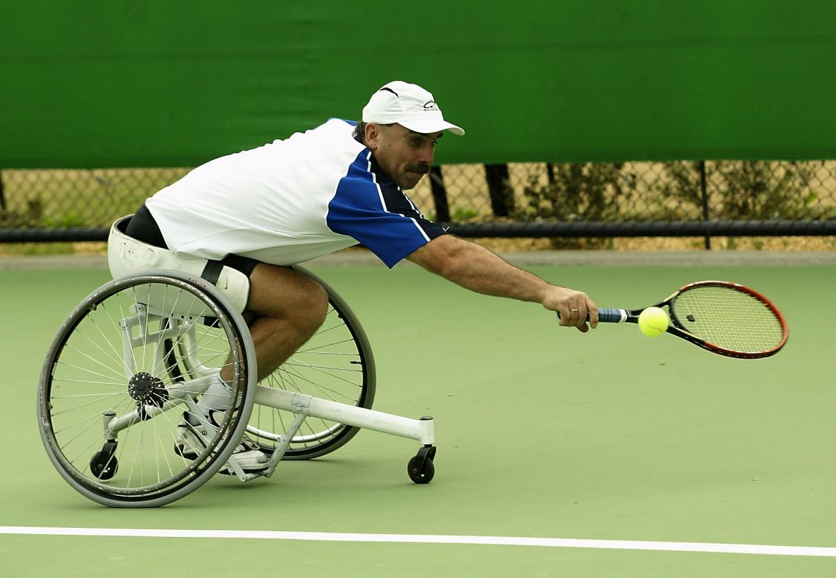 Man in wheelchair reaches to try and hit a tennis ball with his racquet 