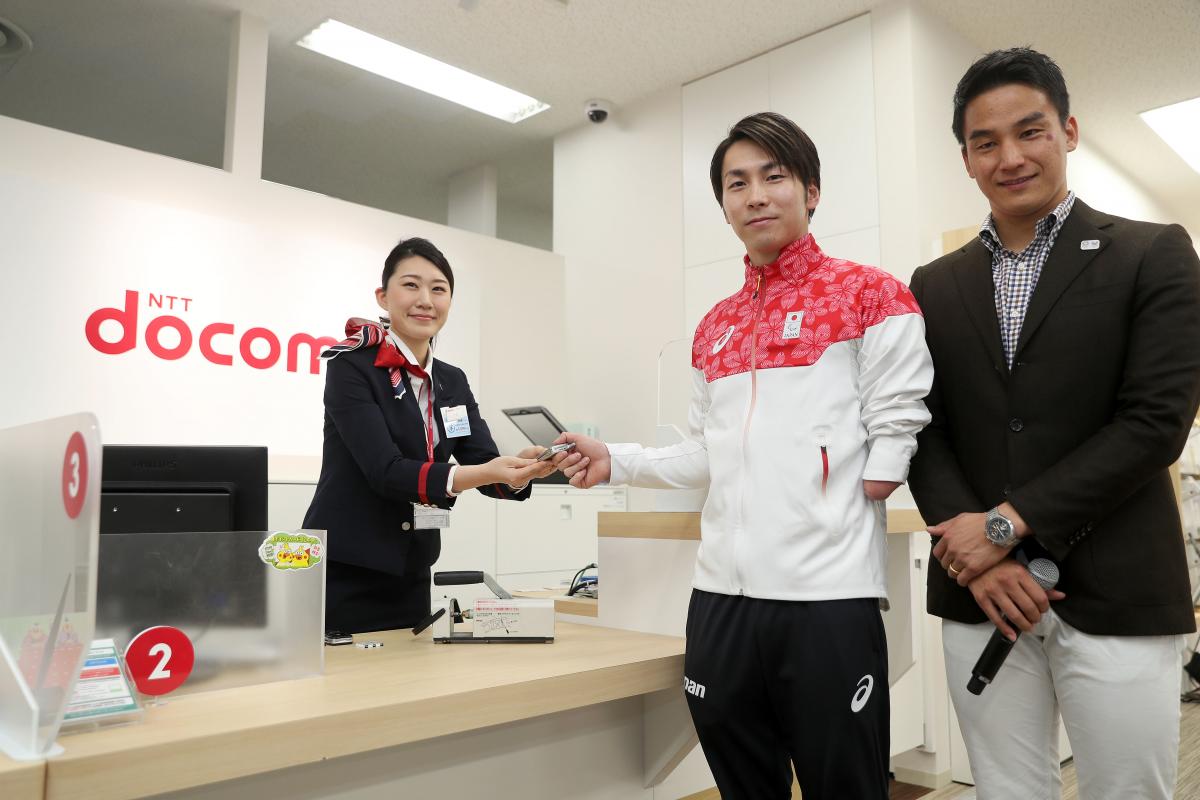 Two Japanese men pose for a photo as a woman hands over a cell phone