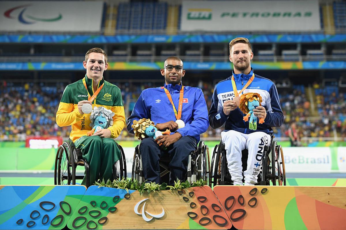 three men in wheelchairs smile holding their medals on the podium